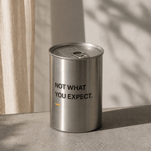 Silver cylindrical scented candle with metal lid, “NOT WHAT YOU EXPECT.” printed
