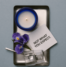 Blue glass OBJECT 05 scented candle in square metal tray with purple flowers and glass holder.