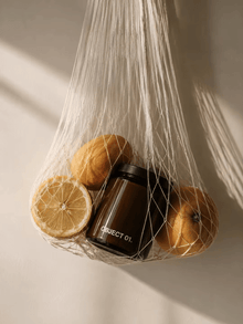 Dark amber glass scented candle jar in mesh bag with citrus.