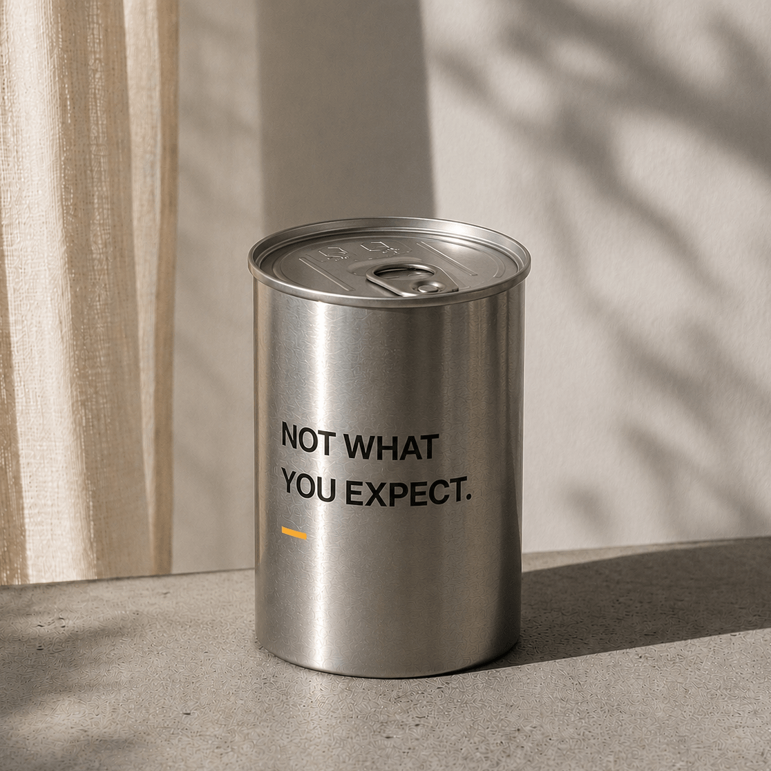 Silver cylindrical scented candle with metal lid, “NOT WHAT YOU EXPECT.” printed