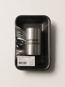 Silver tin scented candle with dark outer sleeve, “NOT WHAT YOU EXPECT.” on label