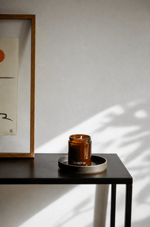 Amber glass scented candle jar on black metal tray in soft sunlight