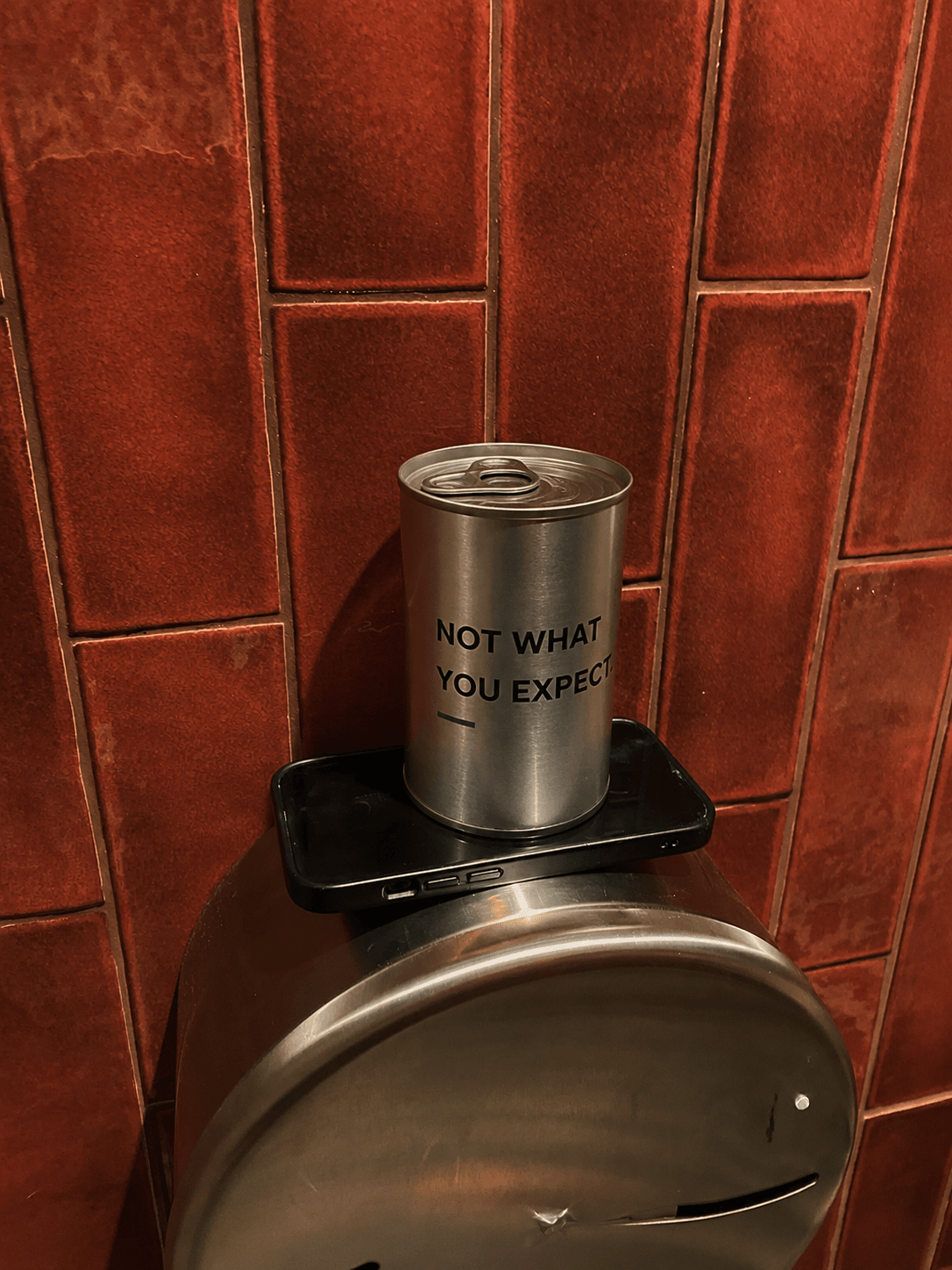 Metal scented candle tin on black holder with label "NOT WHAT YOU EXPECT."