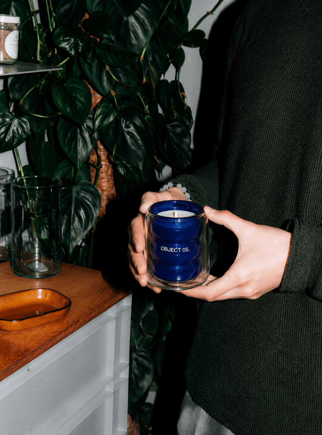 Hands holding a blue OBJECT 05 scented candle jar beside green indoor plants.