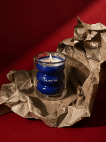 Lit blue glass OBJECT 05 scented candle nestled in crumpled kraft paper packaging.