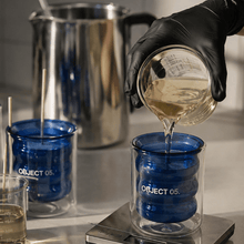 Blue glass candle jar labeled OBJECT 05 with hot wax being poured by a gloved hand