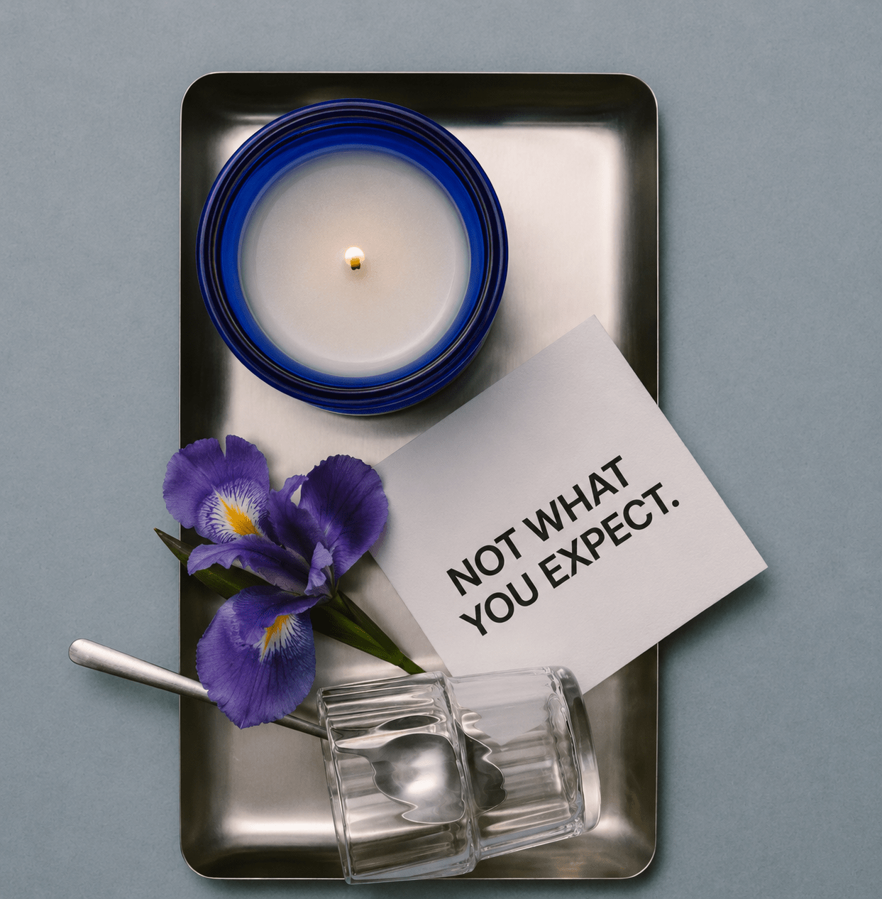 Blue glass OBJECT 05 scented candle in square metal tray with purple flowers and glass holder.
